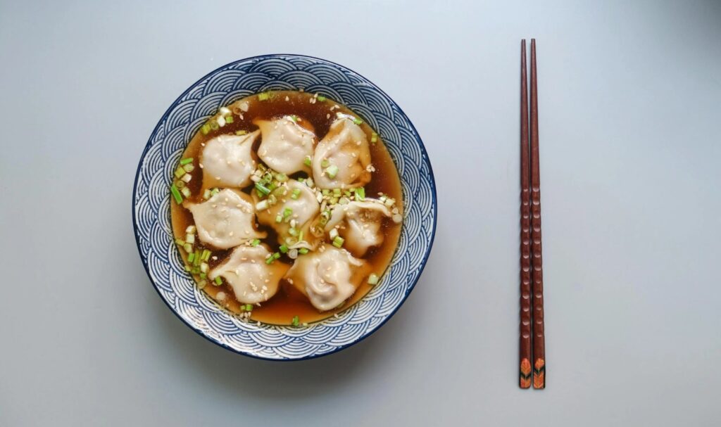 Dumplings in een blauwe kom met bouillon, bosui en sesamzaadjes, geserveerd met houten eetstokjes